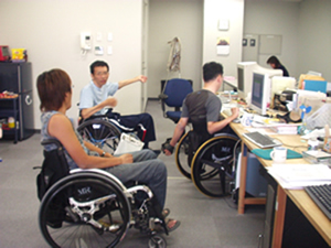複数の人が働くパソコンが並ぶ事務所の中で、１人の車いすの男性と話をしている篠原さんの様子