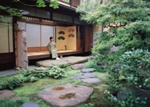 うぐいす色の着物を着ている女性が縁側に座っている日本庭園の風景