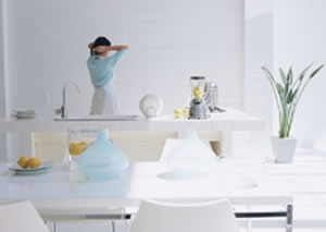 流し台の前でエプロンをつけた女性が髪を束ねていて、手前に白いテーブルがある白い清潔感のあるキッチンの風景